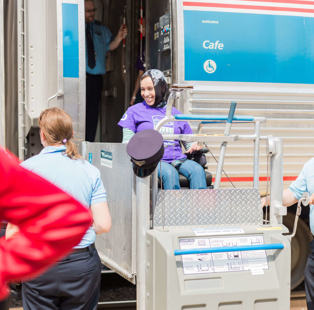 Young woman in headscarf smiles as she uses a wheelchair lift to get off of a train. 