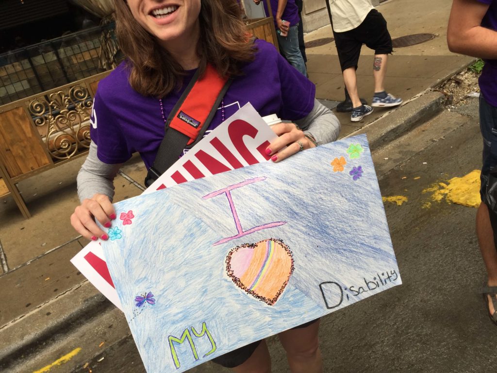 A smiling person with long brown hair carries a sign reading "I heart my disability."