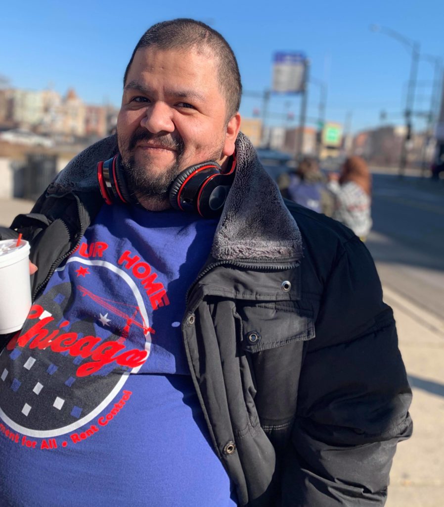 A Latinx man with a disability smiling at the camera. He wears a black winter jacket and a Chicago Cubs shirt.