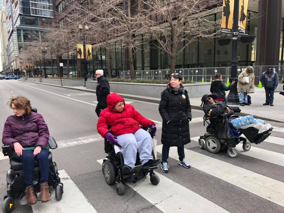 Three people in wheelchairs and one woman who is standing block the street.