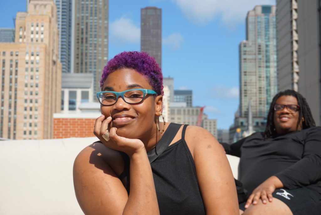 A young black woman with purple hair smiles at the camera.
