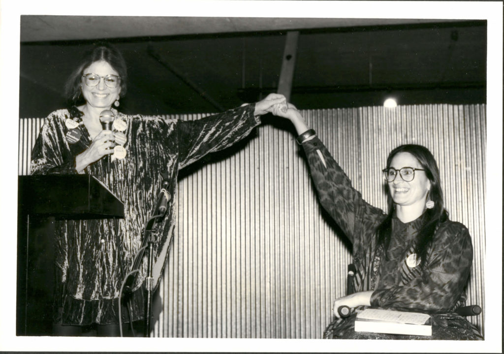 Black and white photo of Gloria Steinem and Marca Bristo, raising clasped hands overhead.