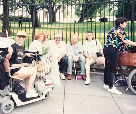 Image is of six disability advocates sitting on a bench in front of the White House in demonstration for the ADA.