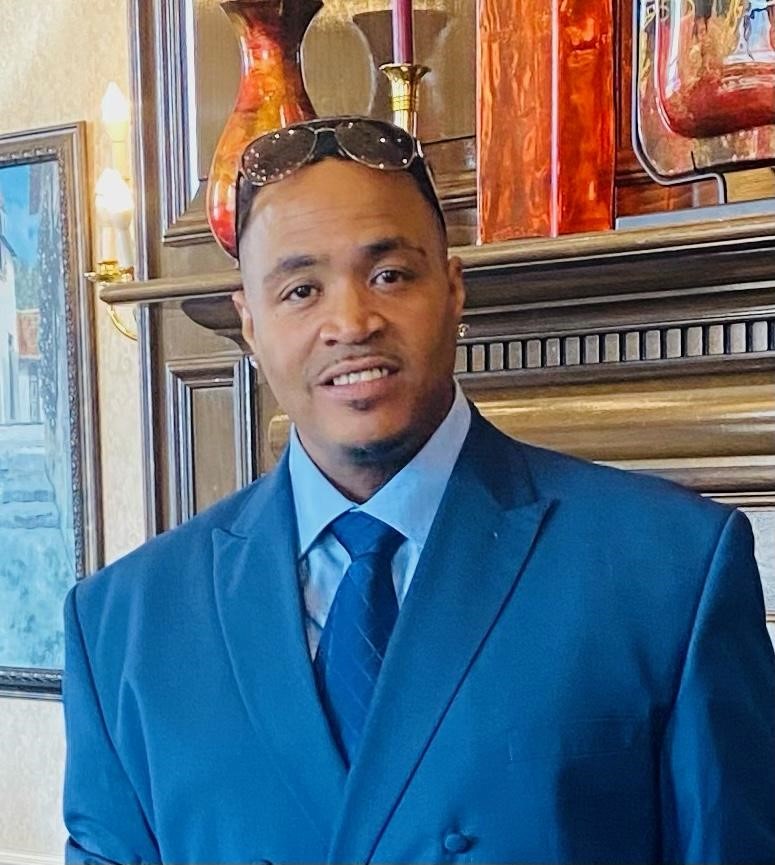 Cedrick Frison, a Black man standing in front of a fireplace mantle. He is wearing a navy blue suit and sunglasses pushed to the top of his head. 