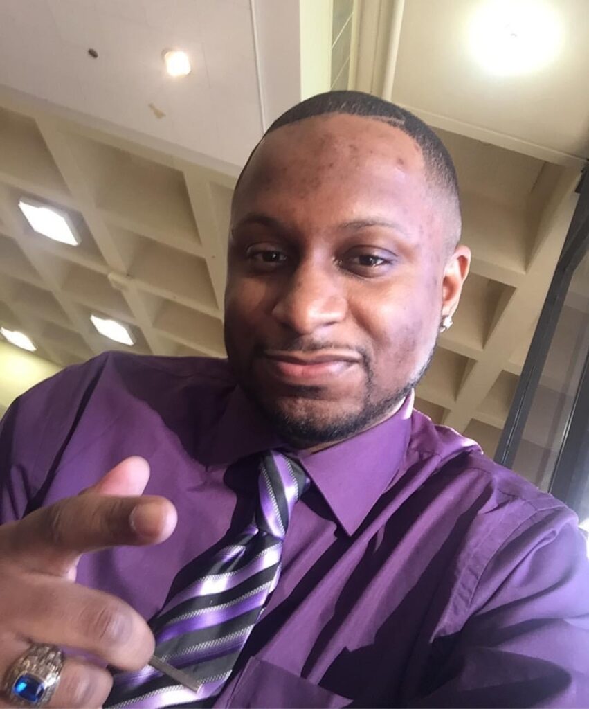 Willie Rolling Jr., a black man wearing a dark purple shirt, a striped tie, and a class ring with a blue stone. He is gesturing towards the camera and smiling. 