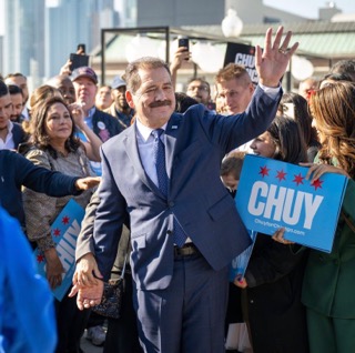 Chuy Garcia, a middle-aged man wearing a suit and tie, waves at a crowd holding campaign signs with his name on them. 