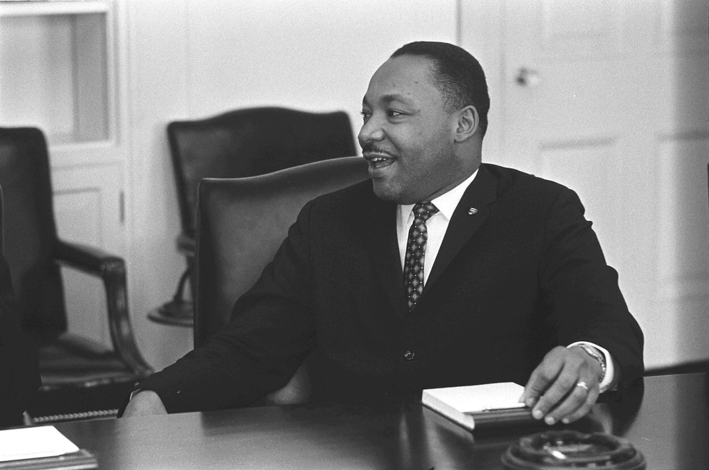 Candid black and white photo of Martin Luther King Jr. smiling and talking to someone seated to his right.