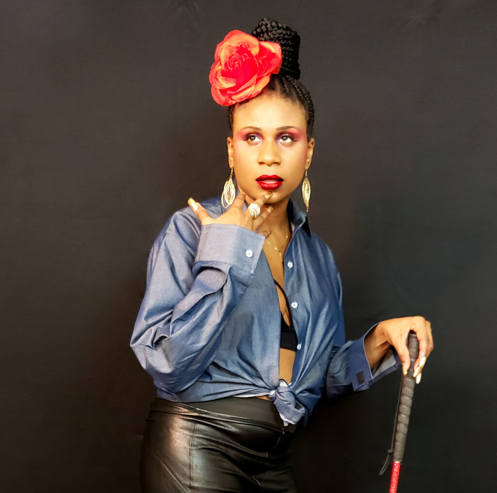 Lachi, a Black woman using a white cane, posing in front of a black backdrop. She has a bright red flower pinned to a braided bun that matches her red lipstick, and is wearing black leather pants and an oxford shirt tied at the waist, exposing her midriff. 