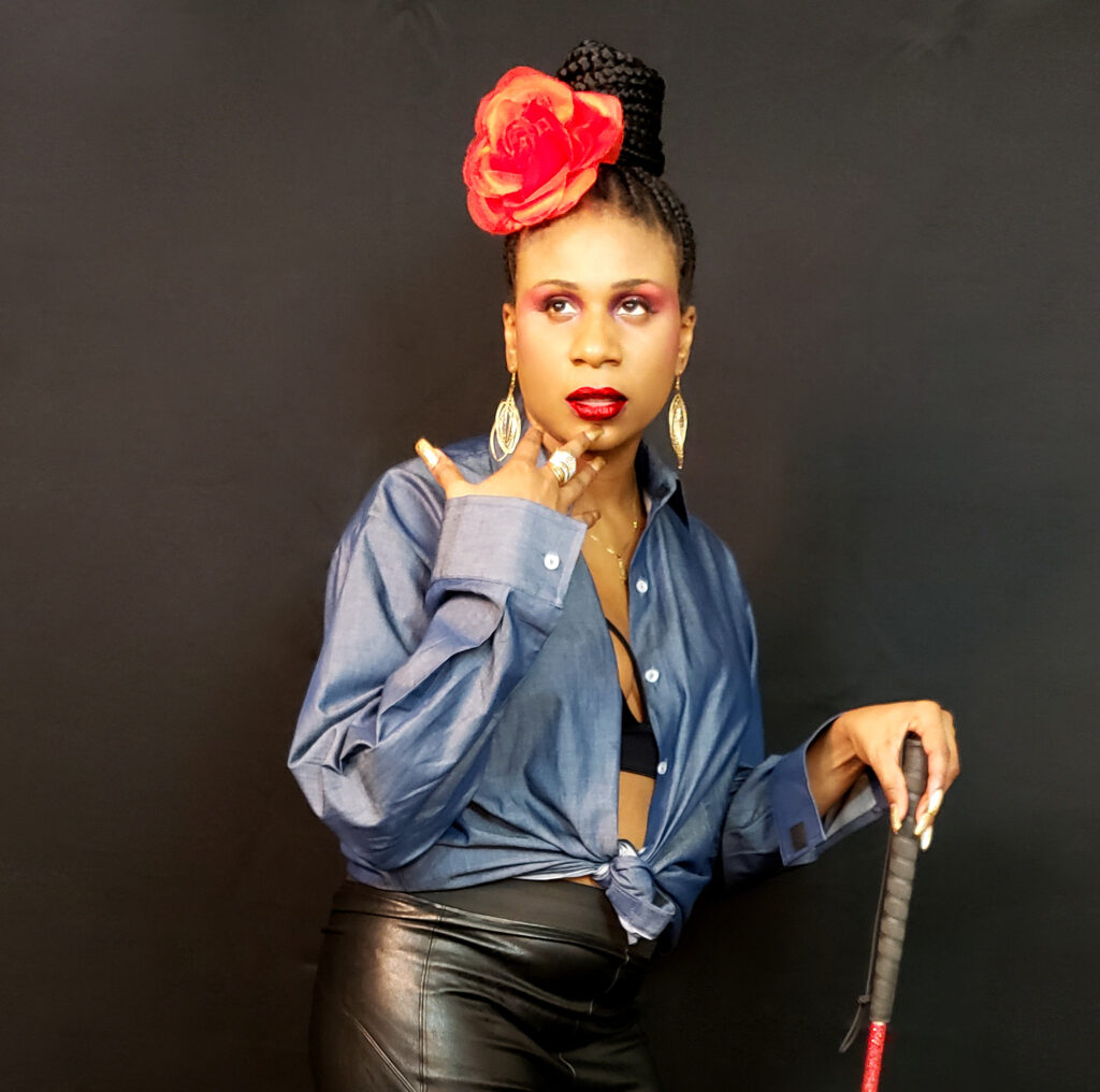 Lachi, a young Black woman, looks off camera, one hand near her lips. She has a bright red flower topping a bun of braids that matches her lipstick. She is wearing dangly earrings, a cambry shirt tied at her waist, and some truly fantastic leather pants. 