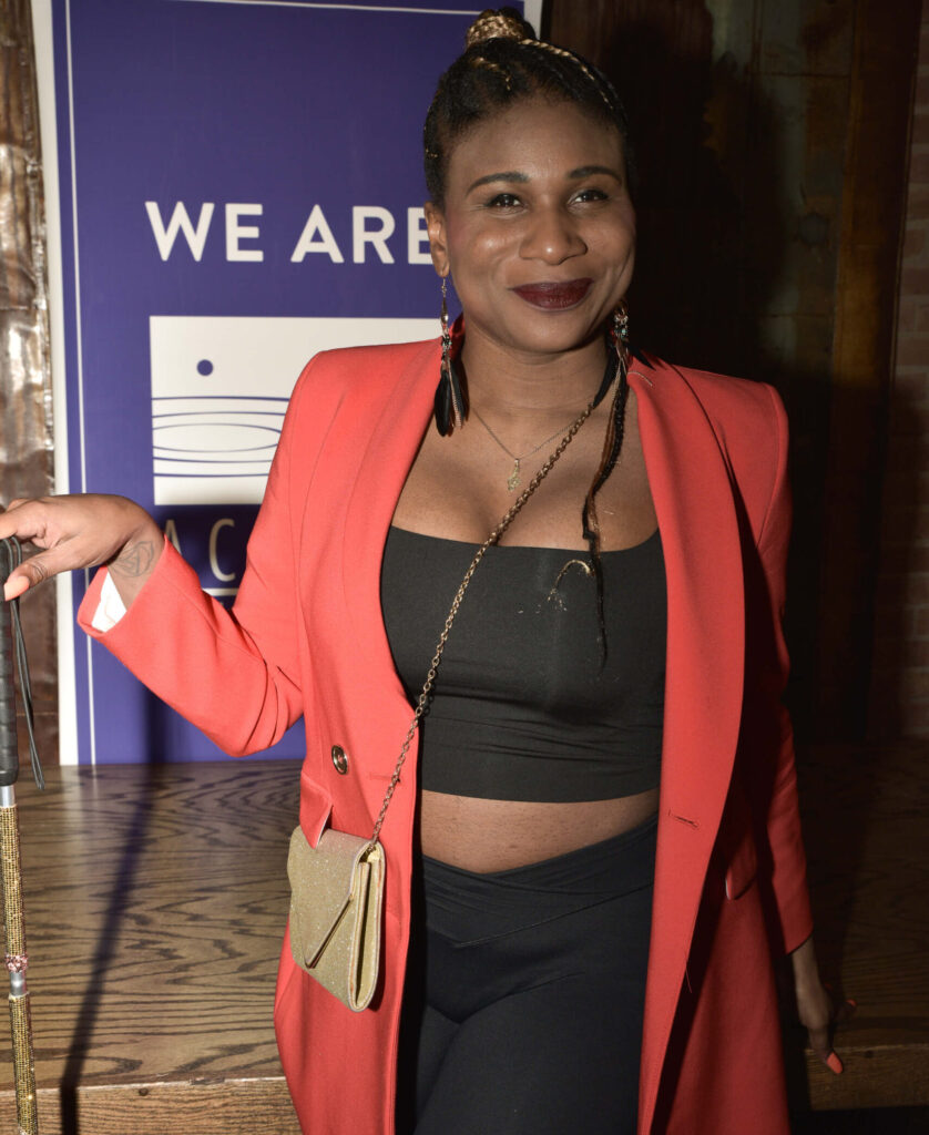 Gala honoree Lachi poses for a picture in front of a purple Access Living banner. She is wearing a black crop top underneath a a bright red jacket and has a sparkly gold handbag.