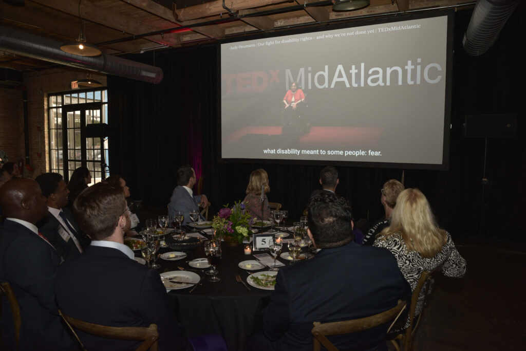 Audience members watching a video tribute to Judy Heumann on a projector screen, which is showing Judy giving a Ted Talk at TedX MidAtlantic. The captions at the bottom read, "...what disability meant to some people: fear."