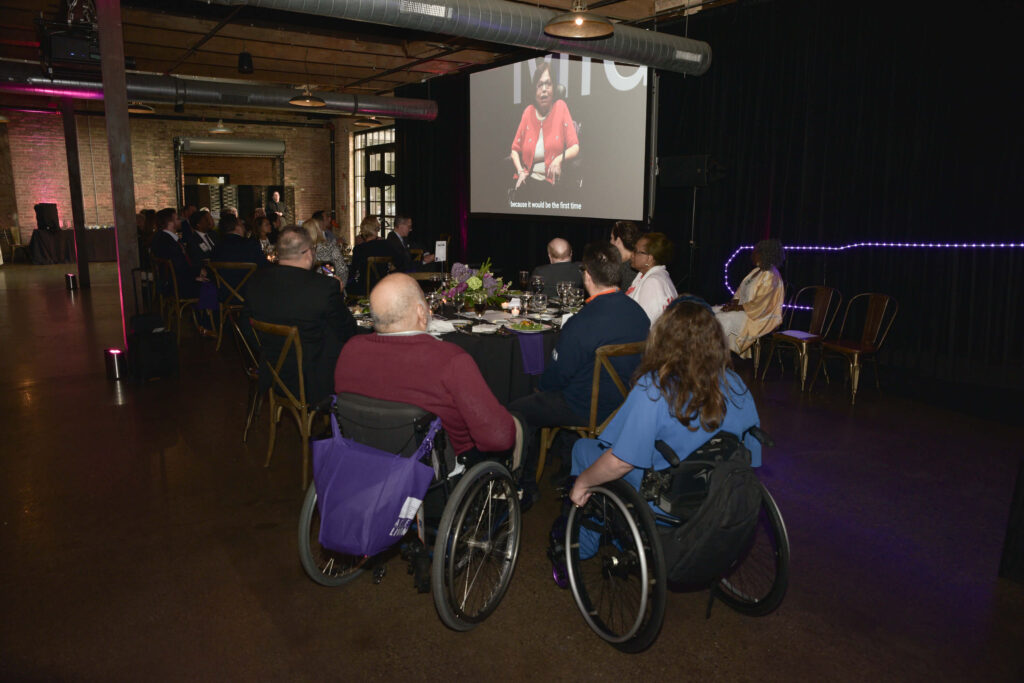 A rear shot of guests watching a video tribute to Judy Heumann. The camera's focus is on Judy's widower, honored guest Jorge Pineda. He watches the video of his late wife speaking from his place in the audience.