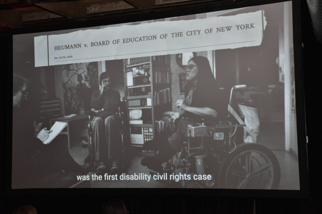 The projector screen as it shows a picture of a young Judy Heumann speaking to several people in a living room. Text across the top of the picture reads, "Heumann v. Board of Education of the City of New York." The captions at the bottom read, "...the first disability civil rights case."