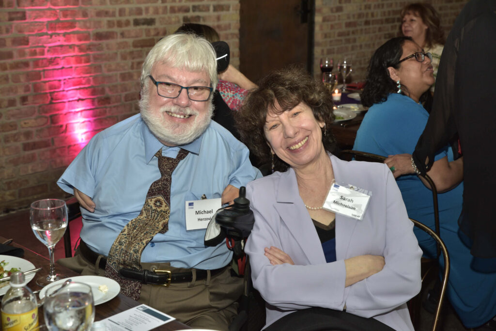 Michael Herzovi and Sarah Michaelson, seated at a table during dinner, smile for the camera.