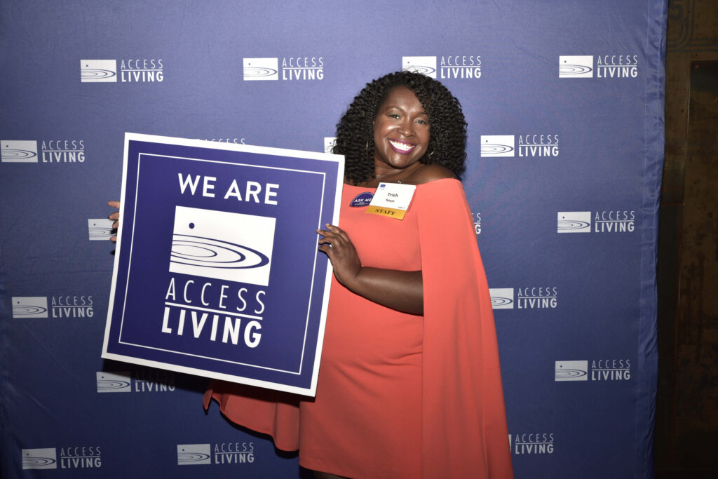 Access Living Director of Independent Living Latricia Seye, holding up a square purple sign that says We Are Access Living. She is standing in front of a purple backdrop with a white Access Living logo repeatedly stamped across it.