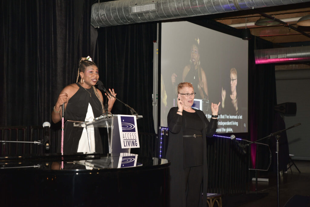 Gala honoree Lachi speaks at a podium on stage with an ASL interpreter to her left.