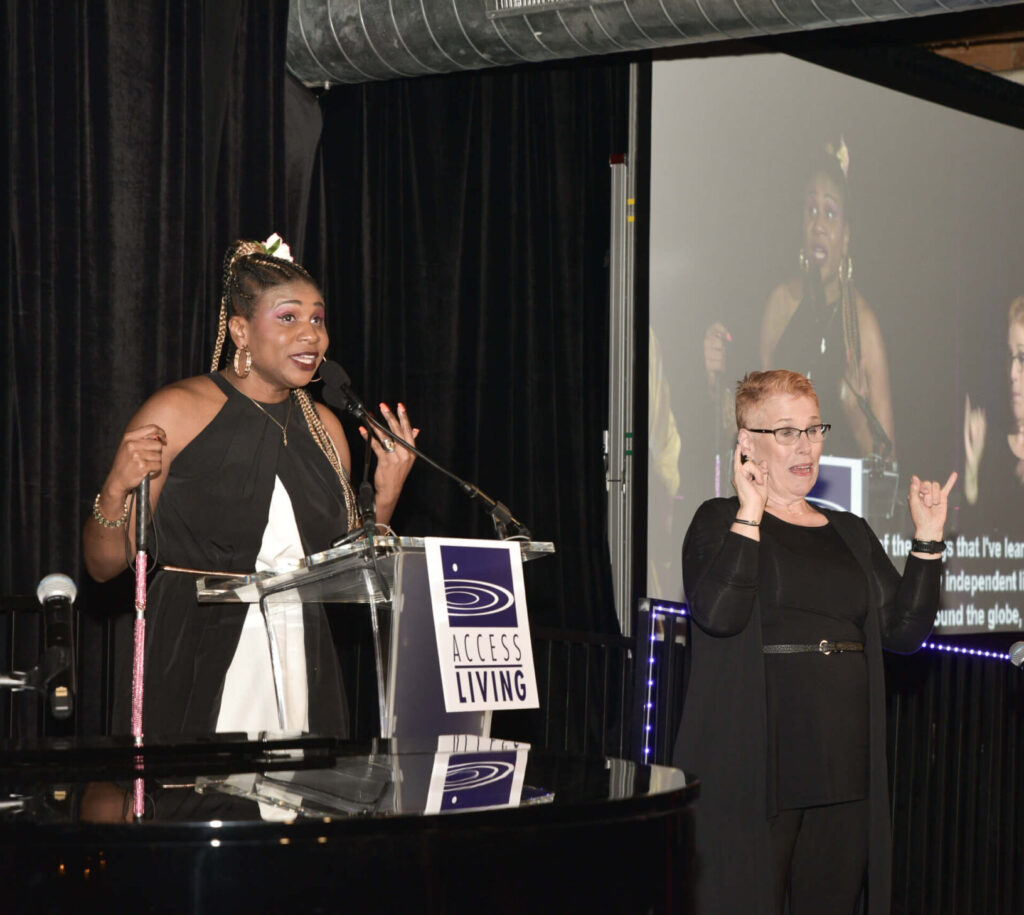 Gala honoree Lachi speaks at a podium on stage with an ASL interpreter to her left.