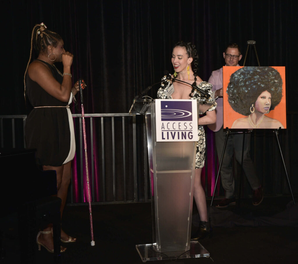 Artist Genevieve Ramos presents gala honoree Lachi with a portrait of herself. Lachi has one hand near her mouth as though in surprised delight. In the portrait on an orange background, a Black woman with a beautiful Afro gazes off to the left. She wears heavy gold earrings and has gold necklaces around her neck. The woman's shoulders are bare.