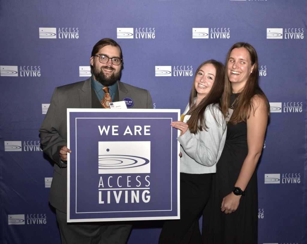 Three members of Access Living's staff (Jerome Palliser, Nichole Shields, and Ashley Eisenmenger) hold up a square purple sign that says We Are Access Living. They're standing in front of a purple backdrop with a white Access Living logo repeatedly stamped across it. They are all grinning.