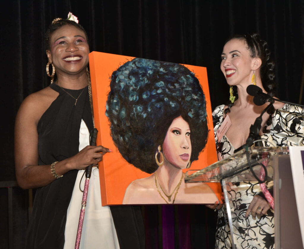 Lachi and artist Genevieve Ramos hold up Genevieve's painting of Lachi together to show the audience. Lachi has tears in her eyes. Both are smiling happily.