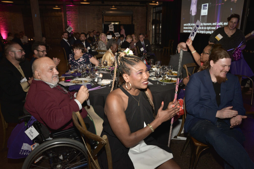 A table of attendees all smiling and clapping. The camera's focus is on gala honoree Lachi as she smiles and applauds.