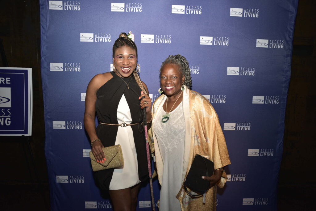 Honoree Lachi poses with the Gala emcee Sylvia Ewing in front of a purple and white Access Living backdrop.