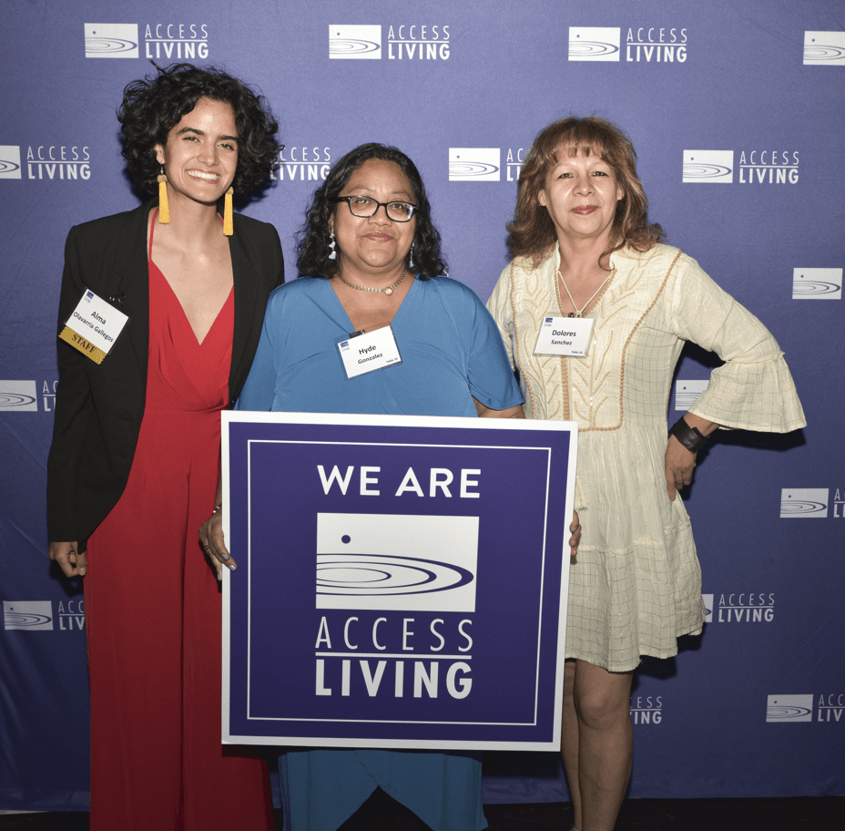 Three women hold up a square purple sign that says We Are Access Living. They're standing in front of a purple backdrop with a white Access Living logo repeatedly stamped across it.