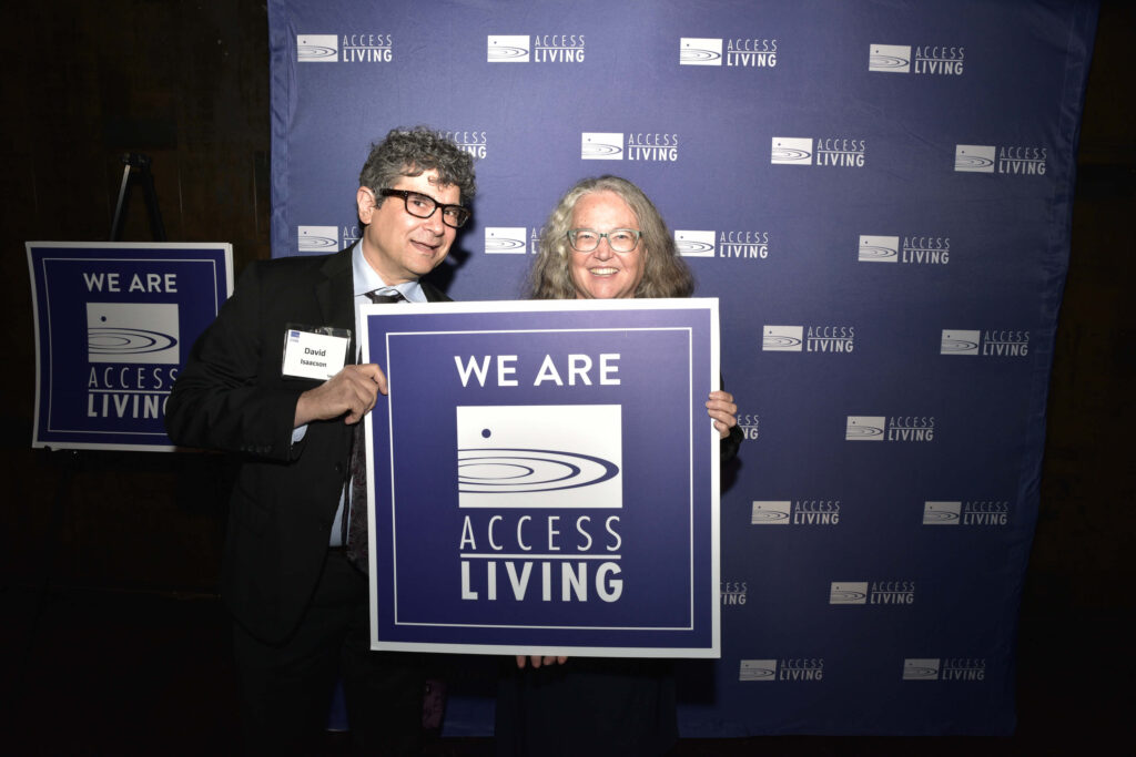 A man and woman hold up a square purple sign that says We Are Access Living. They're standing in front of a purple backdrop with a white Access Living logo repeatedly stamped across it.