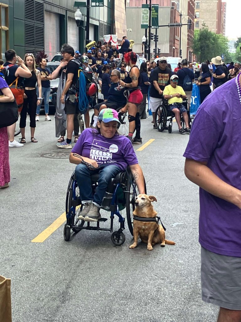 A woman using a wheelchair smiles at the camera, one hand resting on her small brown dog.