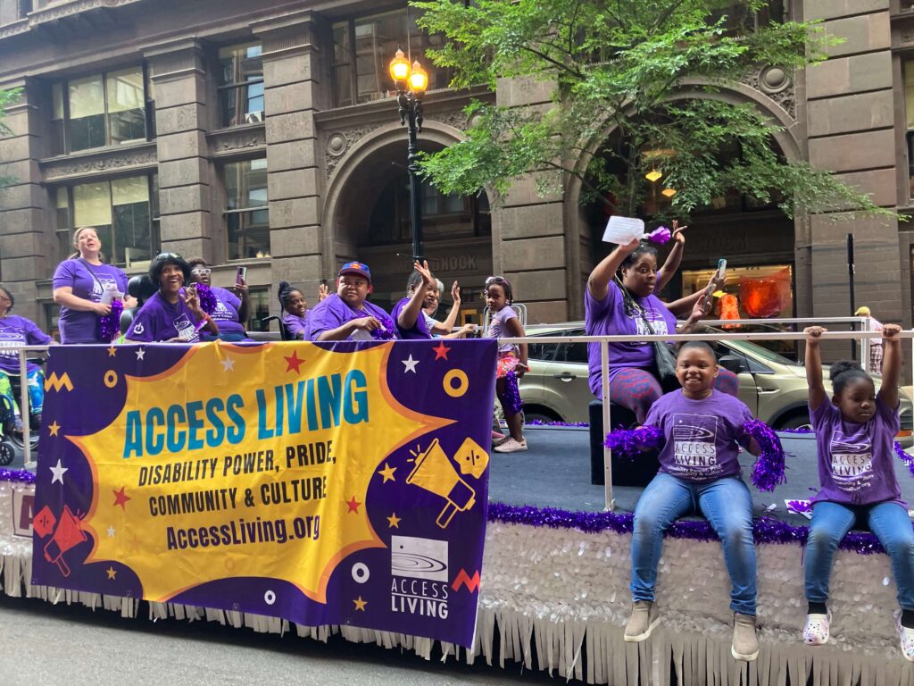 A wide shot of the Access Living parade float. Several adults and a few children wave and smile from the float. On one side is a big sign that reads "Access Living Disability Power, Pride, Community, and CUlture. AccessLiving.org."
