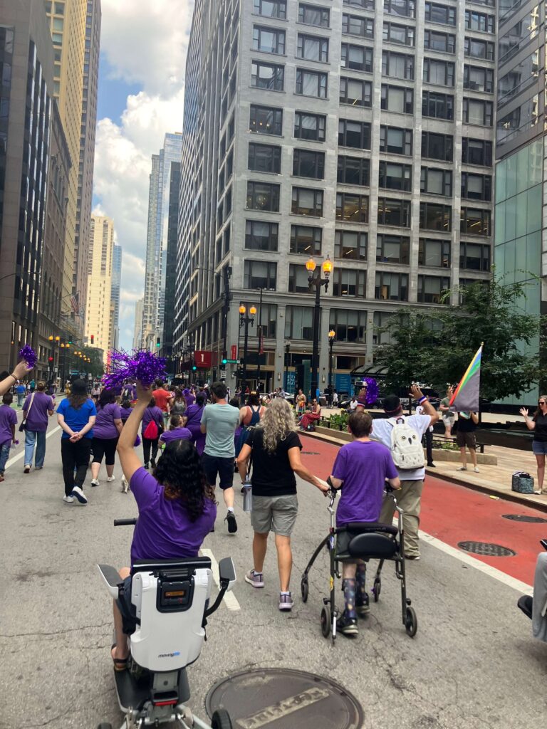 Parade participants cheering as they march down the street. In the foreground is a woman using a scooter and someone else using a walker.
