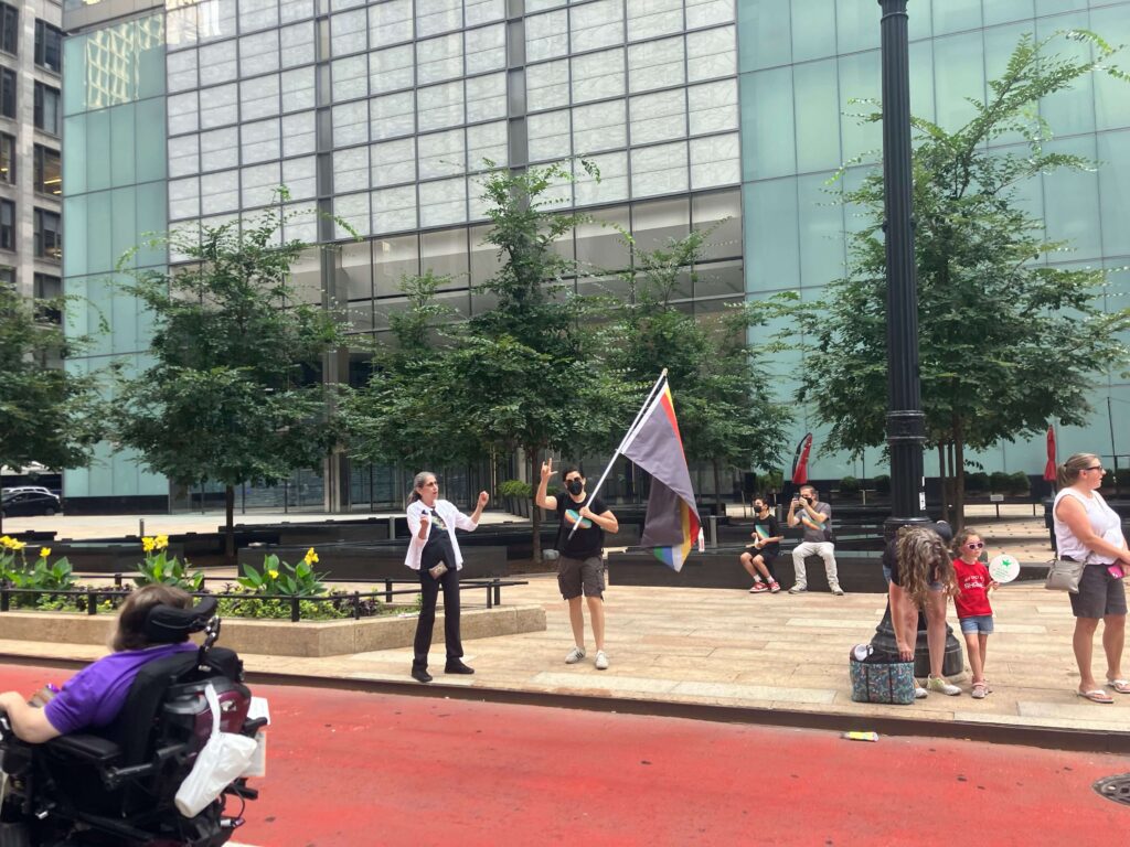 An ASL interpreter signing off to one side. A man carrying the disability rights flag and a face mask looks at the camera, the ASL sign for "love" held over his head