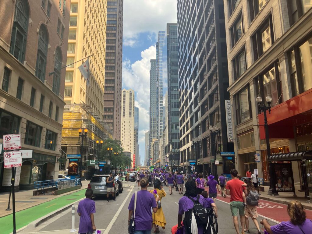 Parade participants walk and roll down an urban street lined with skyscrapers.