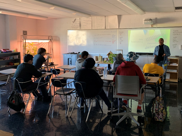 Kids sitting at their desks in a classroom, listening to their teacher.